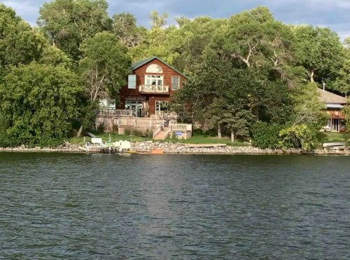A lakeside house surrounded by trees, with a deck and boats visible along the shore.