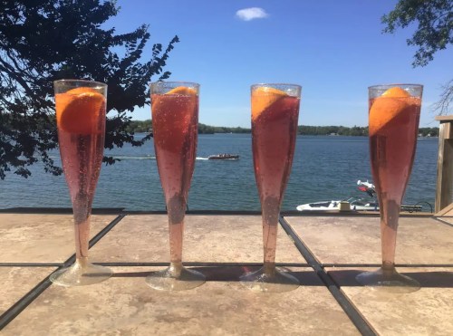 Four sparkling drinks with orange slices sit on a table by a lake, with boats visible in the background under a clear sky.