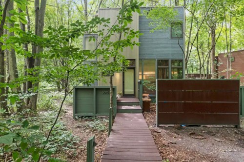 Modern house nestled in a wooded area, featuring large windows and a wooden pathway leading to the entrance.
