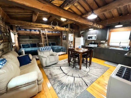 Cozy cabin interior with wooden beams, a dining table, sofa, and two beds in a rustic setting.