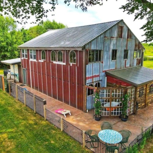 A rustic barn with a metal roof and colorful siding, surrounded by greenery and outdoor seating.