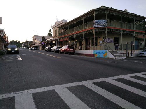 A quiet street scene featuring shops and parked cars, with a crosswalk in the foreground and a welcoming mural on a building.