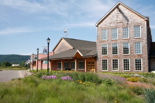 A large building with wooden shingles and multiple windows, surrounded by colorful flowers and greenery under a blue sky.