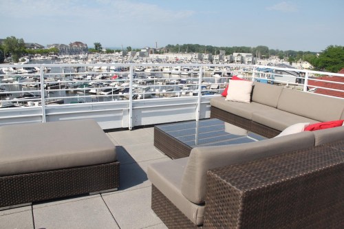Rooftop lounge area with a sectional sofa and glass table, overlooking a marina filled with boats and a clear blue sky.