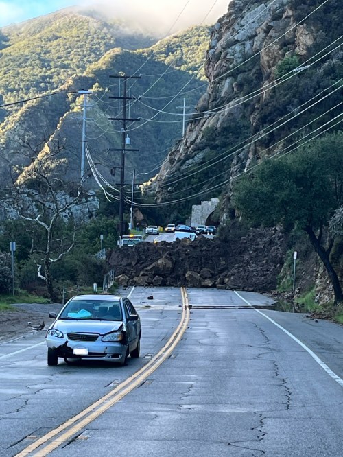 A car is parked on a road blocked by a large landslide, with mountains and power lines in the background.