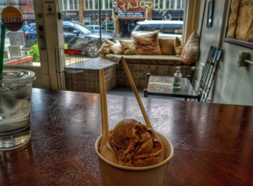 A cup of ice cream with two wooden spoons sits on a table, with a cozy seating area visible in the background.