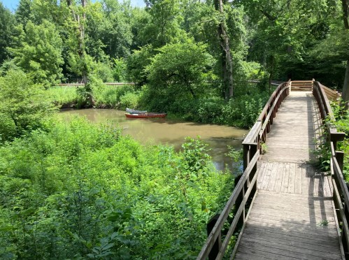 A wooden bridge leads to a serene, overgrown riverbank with a small boat on the water, surrounded by lush greenery.