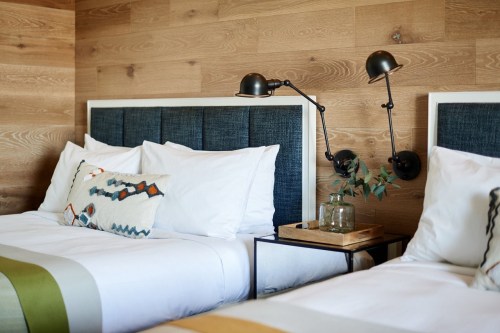 Cozy hotel room with two beds, wooden walls, stylish lamps, and decorative pillows on a nightstand.