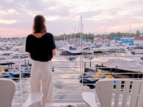 A woman stands on a balcony overlooking a marina filled with boats at sunset.