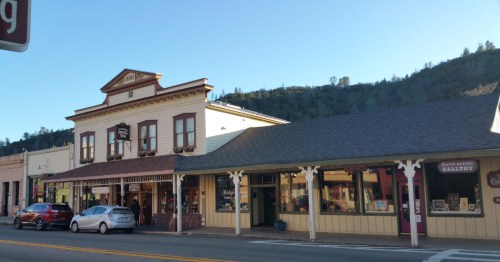 Historic buildings line a street, featuring shops and galleries, with a hillside in the background under a clear blue sky.