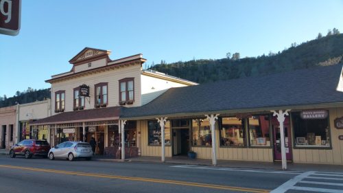 Historic buildings line a street, featuring shops and a gallery, with a mountain backdrop under a clear blue sky.