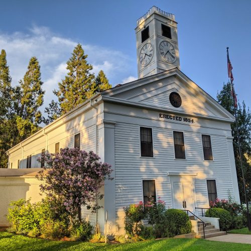 Historic building with a clock tower, surrounded by trees and flowers, under a clear blue sky. Erected in 1894.
