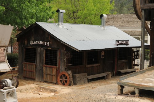 A rustic blacksmith shop with a metal roof, wooden walls, and large windows, surrounded by greenery and gravel paths.