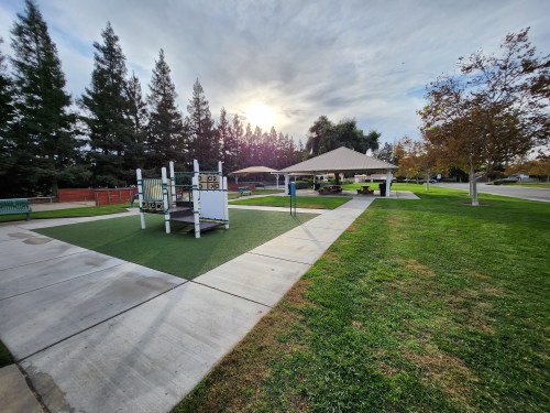 A park scene featuring a playground, picnic area, and trees under a cloudy sky with the sun peeking through.