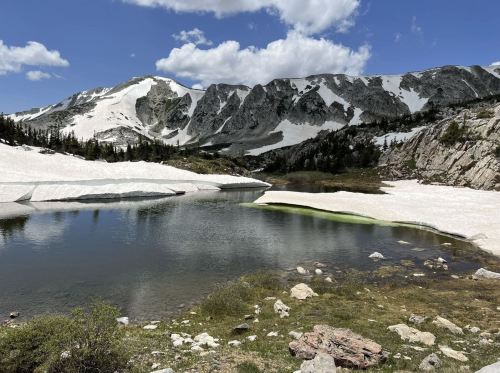 A serene mountain landscape featuring a clear lake, snow-capped peaks, and patches of green vegetation under a blue sky.