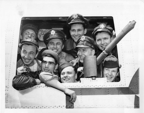 A group of smiling military pilots in uniforms, crowded together in a cockpit window, showcasing camaraderie and joy.