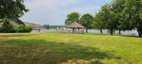 A grassy area by a river, featuring a gazebo and trees under a partly cloudy sky.