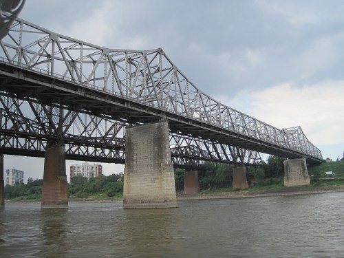 A large metal bridge spans a river, supported by concrete pillars, under a cloudy sky.