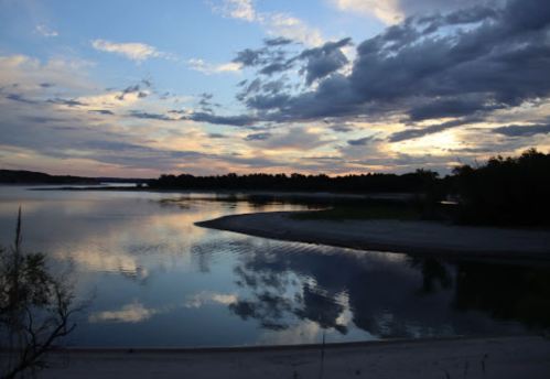 A serene river at sunset, reflecting clouds and trees, with a sandy shore in the foreground.