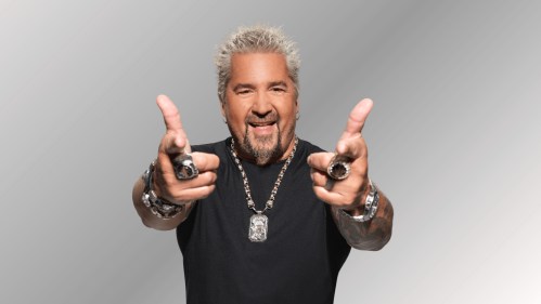 A smiling man with spiky gray hair points playfully at the camera, wearing a black shirt and multiple rings.