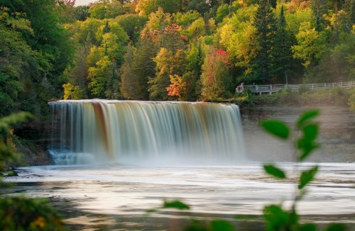 A serene waterfall cascades into a river, surrounded by vibrant autumn foliage and lush greenery.