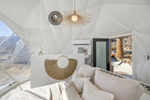 Cozy interior of a geodesic dome with a light fixture, soft furnishings, and large windows showcasing a natural landscape.