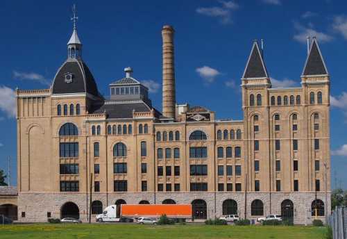 Historic brick building with towers and a smokestack, set against a blue sky and green grass.