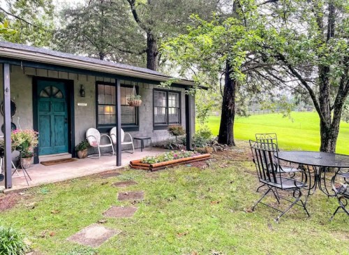 A cozy cottage with a teal door, surrounded by greenery and a small patio set on a grassy lawn.