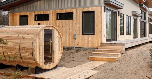 A wooden sauna barrel next to a modern house with large windows and a wooden deck.