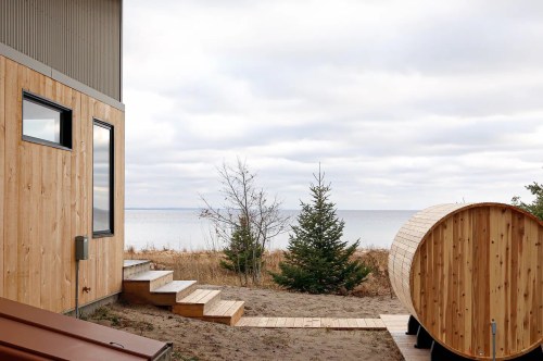 A wooden cabin by the water, featuring steps leading to a sauna and a view of the lake under a cloudy sky.