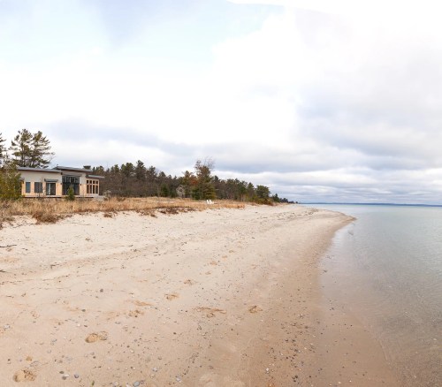 A sandy beach with gentle waves, lined by trees and a modern building in the background under a cloudy sky.