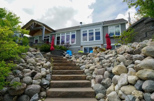 A stone pathway leads up to a house with large windows, surrounded by greenery and red umbrellas on the patio.