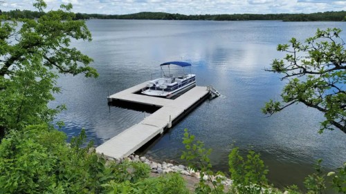 A serene lake view featuring a dock with a boat, surrounded by lush greenery and a cloudy blue sky.