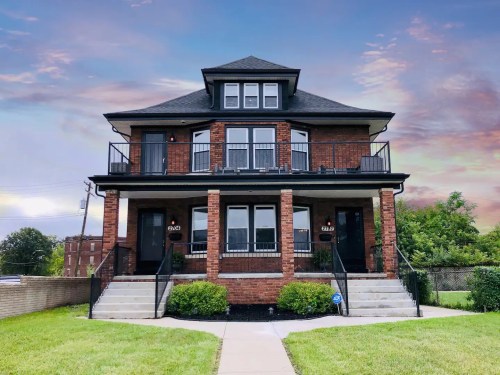 A two-story brick duplex with balconies, surrounded by green grass and a colorful sky at sunset.
