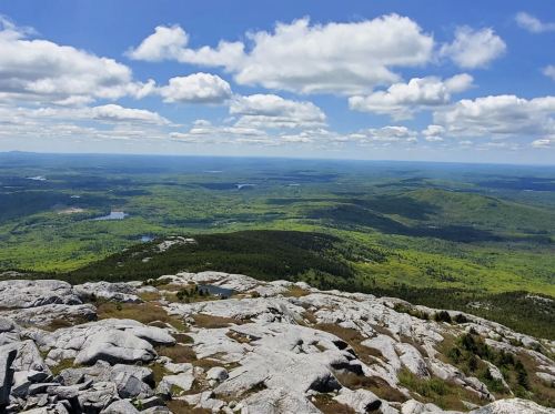 A panoramic view from a rocky mountain summit, showcasing lush green valleys and a blue sky with fluffy clouds.