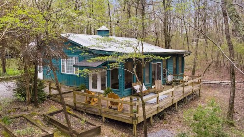 A cozy blue cabin surrounded by trees, featuring a wooden deck and outdoor seating.