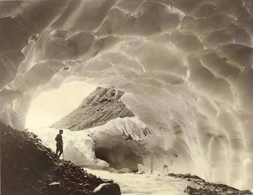 A person stands inside a glacial cave, surrounded by ice formations and a mountain in the background.