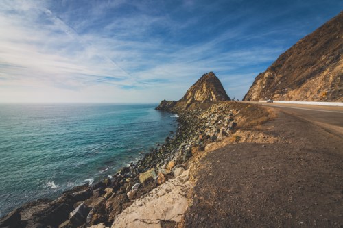 A scenic coastal view with rocky cliffs, clear blue water, and a winding road along the shoreline under a blue sky.