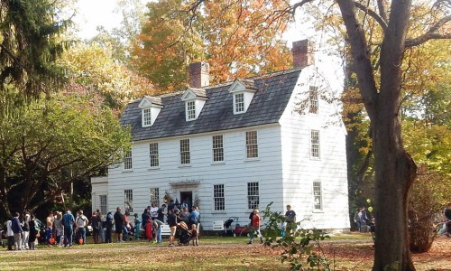 A historic white house surrounded by trees, with a crowd of people gathered outside in autumn.