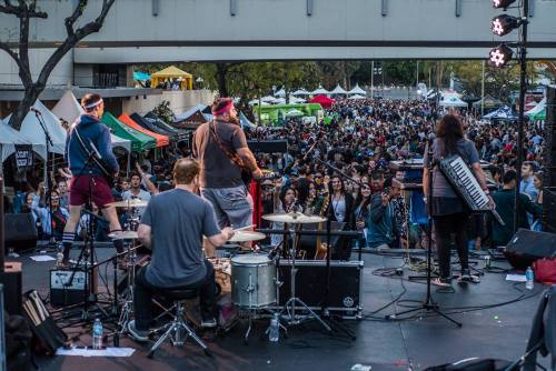 A band performs on stage in front of a large crowd at an outdoor festival, with colorful tents in the background.