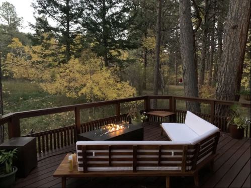 A cozy outdoor deck with a fire pit, surrounded by trees and autumn foliage. A white couch adds to the inviting atmosphere.