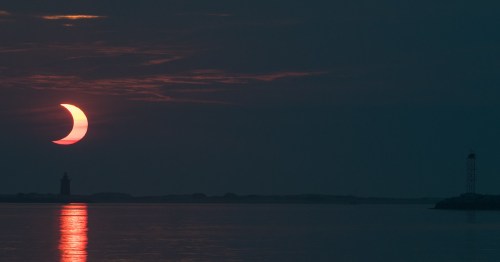 A crescent sun sets over calm waters, reflecting light, with a silhouette of a lighthouse in the distance.