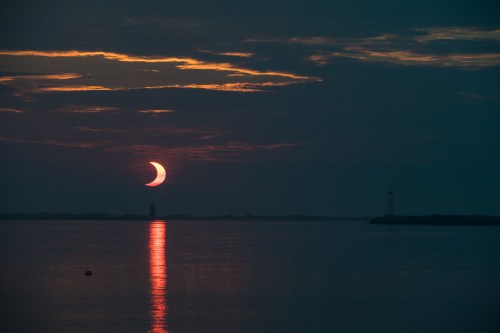 A crescent moon rises over a calm sea, reflecting light on the water with a lighthouse in the distance.