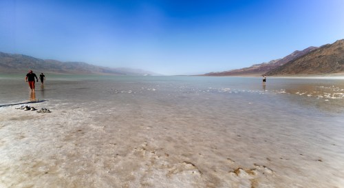 People walking on a salt flat by a calm, shallow lake with mountains in the background under a clear blue sky.