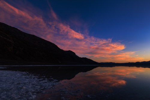 A serene sunset over a calm lake, with vibrant clouds reflecting on the water and mountains in the background.