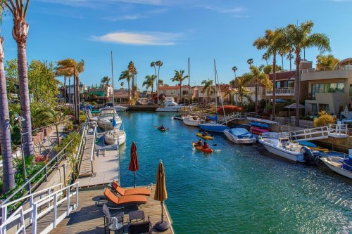 A scenic canal with boats and kayakers, surrounded by palm trees and waterfront homes under a clear blue sky.