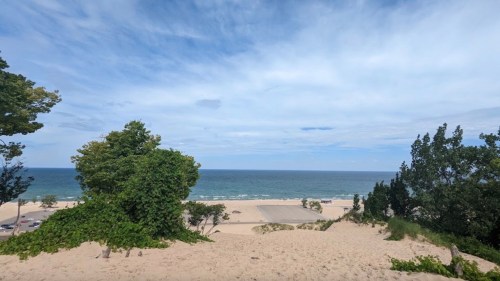 A sandy beach with trees in the foreground and a calm ocean under a partly cloudy sky.