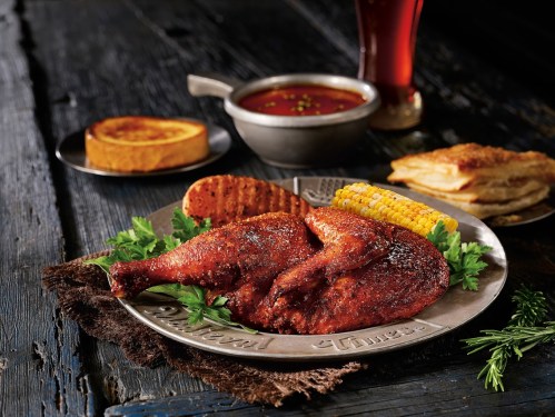 A platter with two roasted chicken legs, corn, a slice of bread, and a bowl of sauce on a rustic wooden table.