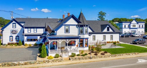 A charming blue and white Victorian-style building with a porch, surrounded by greenery and a parking lot.
