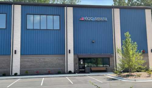 Exterior of a blue and gray building with a sign that reads "KOC AERIAL" and a landscaped parking area.